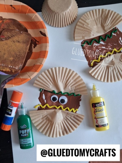 Three craft projects use brown-painted cupcake liners to make cartoon tacos with googly eyes and painted toppings, incorporating fun spatula stamped crafts for added texture. Art supplies like glue sticks, paint, and brushes are nearby. @GLUEDTOMYCRAFTS is labeled on the image.