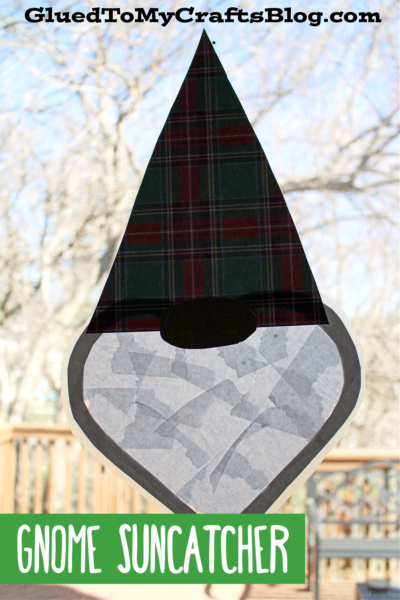 A gnome suncatcher with a plaid hat and gray beard is taped to a window. Trees and a wooden railing can be seen outside. The text GNOME SUNCATCHER appears at the bottom.