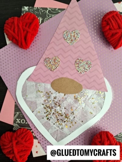 A pink paper gnome suncatcher craft with a heart-shaped beard decorated with tissue paper and glitter, a brown oval nose, and three glitter hearts on the hat. Red yarn hearts and patterned paper in the background. @GLUEDTOMYCRAFTS at the bottom.