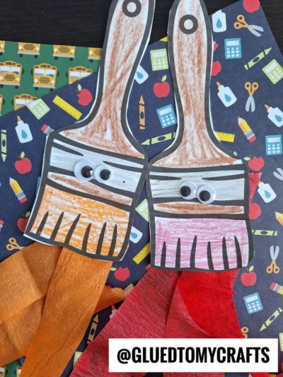 Two paint brush craft projects with googly eyes and orange and red tissue paper bristles are displayed on colorful school-themed backgrounds. A @GLUEDTOMYCRAFTS label is at the bottom.