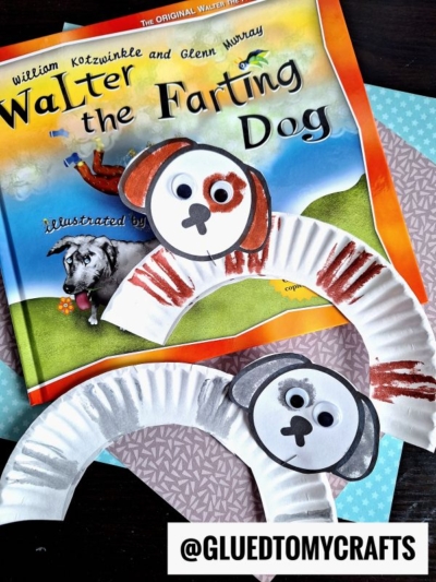 Two paper plate dog crafts—one brown and one gray—are displayed on a book titled Walter the Farting Dog. Featuring large eyes and floppy ears, each paper plate dog sits atop patterned blue paper for a playful touch.