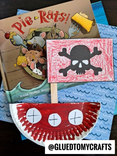 A children’s book titled Pie-Rats! is shown with two crafts: a paper plate pirate ship featuring red and white X marks for a pirate smile, and a red crayon-colored pirate flag with a black skull and crossbones on a stick.