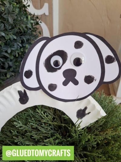 A paper plate dog with a white face, black spots, and googly eyes is attached to a white arch, surrounded by green plants. The handle @GLUEDTOMYCRAFTS appears in a green box at the bottom.