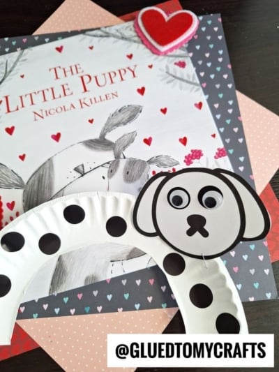A children’s book titled The Little Puppy by Nicola Killen is surrounded by heart-themed paper. A paper plate dog craft with googly eyes and black spots is in the foreground. The handle @GLUEDTOMYCRAFTS is at the bottom.