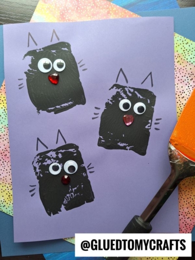 A purple sheet with three black paint cat faces made from spatula stamped crafts and sponge prints, each with googly eyes, red nose, and drawn ears and whiskers. Craft tools and colorful paper lie around. Text: @GLUEDTOMYCRAFTS.