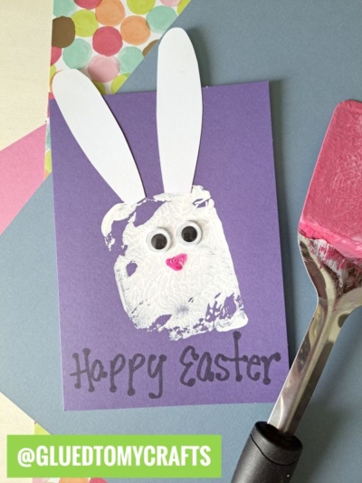 A purple card features a bunny face made from a spatula stamped craft with a white sponge print, googly eyes, a pink heart nose, and white paper ears. Happy Easter is written below. Colorful paper pieces are beside the card.