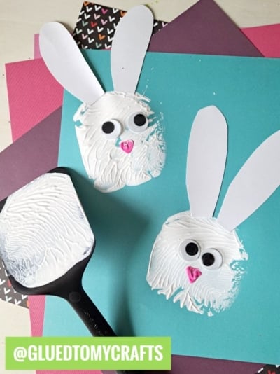 Two bunny faces made from white paint and spatulas, with paper ears, googly eyes, and pink foam noses, are displayed on colorful paper. Perfect for spatula stamped crafts, a spatula with paint and the @GLUEDTOMYCRAFTS handle also appear in the image.