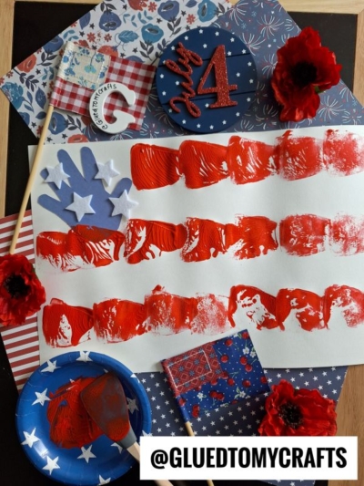 A hand-painted American flag craft with red paint stripes and white handprints for stars, surrounded by red flowers, patterned papers, a paintbrush, and fun spatula stamped crafts accents. @GLUEDTOMYCRAFTS is displayed at the bottom.