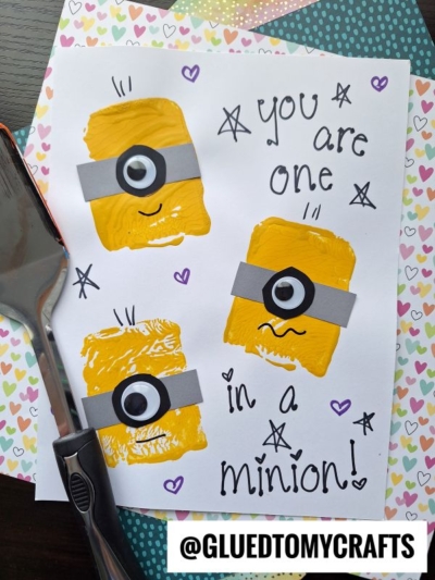 A handmade card features three yellow Minions made from paint and paper, each with different facial expressions. The text reads, You are one in a minion! The card is on heart-patterned paper with spatula stamped crafts beside it.