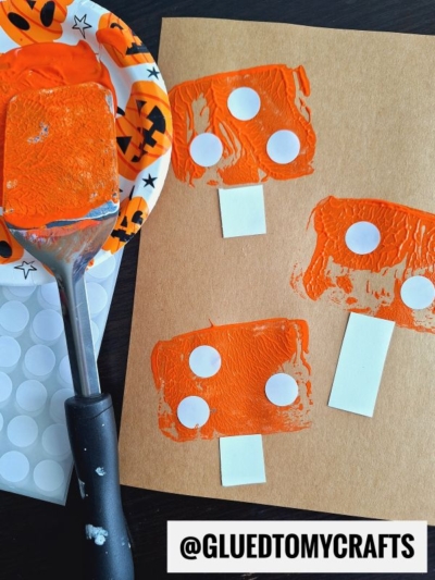 A spatula stamped crafts project featuring orange paint, a Halloween-themed plate, and brown paper with orange mushroom shapes decorated with white stickers. The handle displays the Instagram handle @GLUEDTOMYCRAFTS.