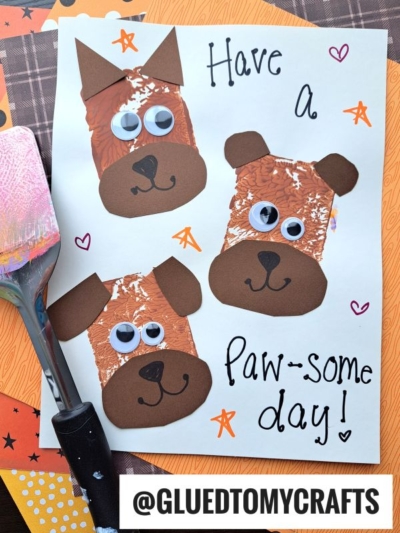 A handmade card with three brown dog faces made from painted paper, googly eyes, and cut-out shapes—an adorable example of spatula stamped crafts. The card says Have a paw-some day! and is surrounded by craft supplies and colorful paper.