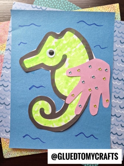 A colorful handprint seahorse craft made with green and yellow paper, a pink handprint cutout as its fin, a googly eye, and blue wavy lines on blue paper. The handle @GLUEDTOMYCRAFTS is at the bottom.