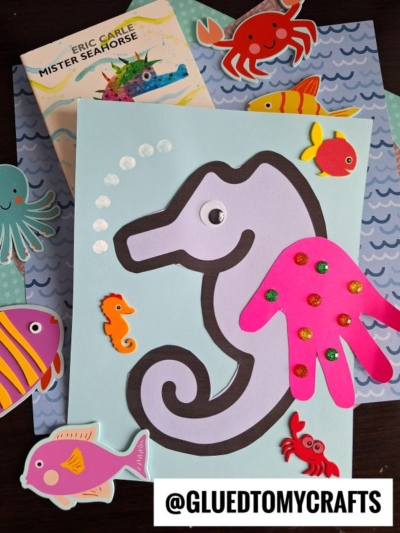 A children’s art project featuring a handprint seahorse made from paper, with a pink handprint as its fin, a googly eye, decorative stickers, colorful sea creatures, and the book Mister Seahorse.
