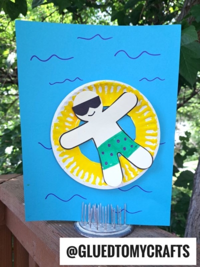 A paper craft of a person in sunglasses and polka-dot shorts floats on a yellow pool floatie art against a blue background with wavy lines, resembling water. The Instagram handle @GLUEDTOMYCRAFTS is at the bottom.