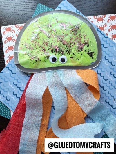 Colorful jellyfish suncatcher craft made from green tissue paper, glitter, googly eyes, and orange, red, and blue streamers for tentacles, displayed on sea-themed patterned paper. @GLUEDTOMYCRAFTS is visible at the bottom.