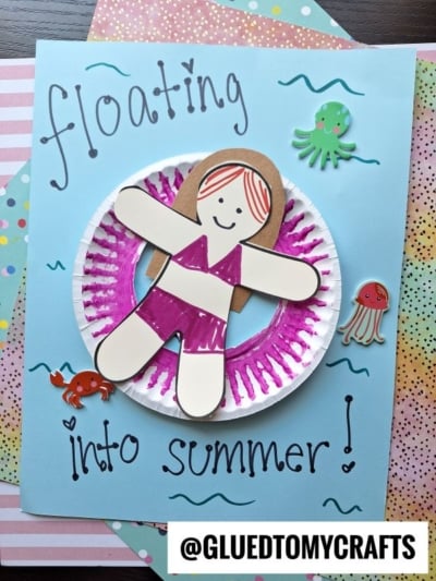 A handmade craft shows a paper girl in a purple swimsuit relaxing on a pool floatie art piece atop a decorated plate, with "floating into summer!" and sea creature stickers on a pastel background.