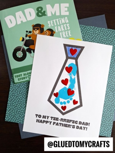 A handmade Fathers Day card with a footprint tie design featuring blue child footprints and red hearts, next to a book titled Dad & Me: Setting Farts Free. The card reads, To my tee-rriffic dad! Happy Fathers Day!.