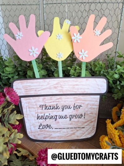 A handmade craft features a paper flower pot with three handprint flowers, decorated with paper daisies. The pot reads, Thank you for helping me grow! Love, _____. Flowers and greenery surround the craft. Text: @GLUEDTOMYCRAFTS.