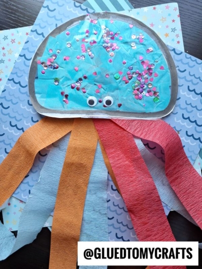 A colorful jellyfish suncatcher craft made with blue paper, tissue paper tentacles in orange, red, and blue, sequins, and googly eyes, displayed on patterned backgrounds. @GLUEDTOMYCRAFTS is written at the bottom.