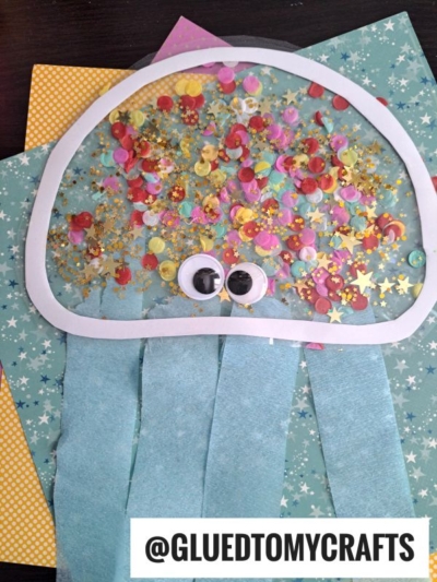 A colorful jellyfish suncatcher craft made from paper, confetti, glitter, and googly eyes on a blue and yellow star-patterned background. The jellyfish features blue paper streamers as tentacles.