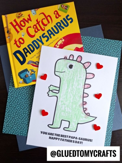 A children’s book titled “How to Catch a Daddysaurus” and a handmade papa-saurus card decorated with red hearts sit on blue paper. The card reads, “You are the best papa-saurus! Happy Father’s Day!”.