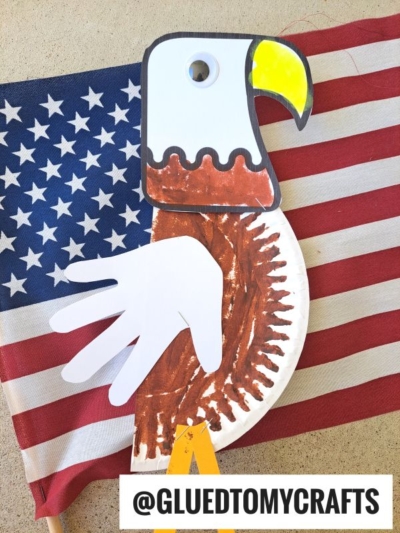 A handmade paper plate bald eagle craft, featuring a cutout hand for its wing, is placed in front of an American flag. The eagle’s head is white with a yellow beak, and its body and wing are brown. @GLUEDTOMYCRAFTS is written at the bottom.