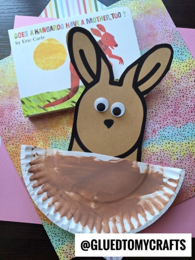 A paper plate kangaroo craft made from brown paper and a painted plate with googly eyes is shown beside the book Does a Kangaroo Have a Mother, Too? by Eric Carle, on colorful paper backgrounds.