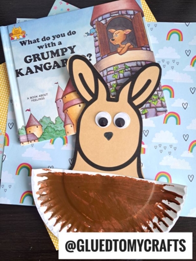 A children’s book titled “What do you do with a Grumpy Kangaroo?” sits on rainbow-patterned paper next to a homemade paper plate kangaroo craft. The handle @GLUEDTOMYCRAFTS is displayed at the bottom.