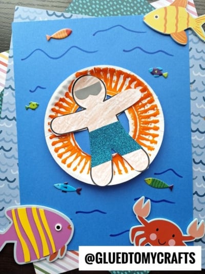 A paper craft of a person with sunglasses in a glittery swimsuit floats on an orange sunburst pool floatie art, surrounded by colorful fish, a crab, and water-themed stickers on a blue background.