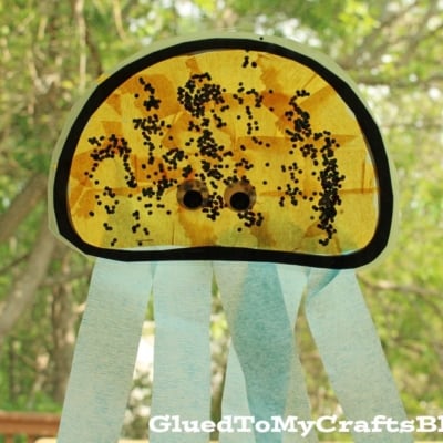 A jellyfish suncatcher craft made from a semicircle coffee filter with black spots, two googly eyes, and blue streamers hanging down, displayed outdoors with green trees in the background.