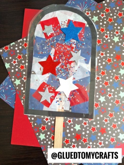 A patriotic popsicle suncatcher craft featuring a popsicle stick handle and a window-shaped frame, decorated with red, white, blue, and silver stars, glitter, and star-patterned paper. The text @GLUEDTOMYCRAFTS is at the bottom.