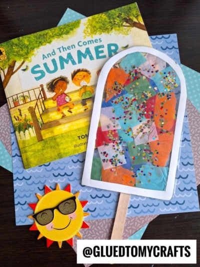 A children’s book titled “And Then Comes Summer” sits next to two crafts: a smiling sun with sunglasses and a colorful popsicle suncatcher made of tissue and glitter. The background features patterned blue paper.