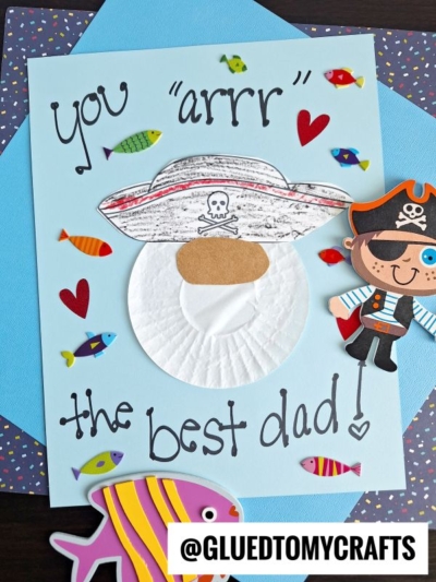A handmade Fathers Day card featuring a cupcake liner pirate face with "You arrr the best dad!" surrounded by fish stickers and hearts. @GLUEDTOMYCRAFTS at the bottom.