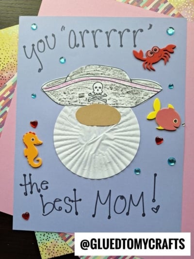 A handmade card showcases a cupcake liner pirate face with a beard, a paper hat topped with a skull, and sea animal stickers. It reads, You arrrrrr the best MOM! The @GLUEDTOMYCRAFTS handle appears at the bottom.