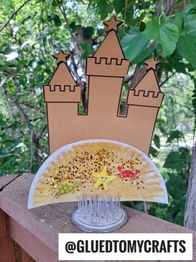 A cardboard and paper plate sand castle with star, turtle, and crab stickers is displayed outdoors on a wooden ledge. Gold glitter decorates the sand, and trees are visible in the background. @GLUEDTOMYCRAFTS is written at the bottom.