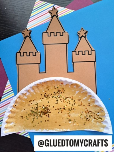 A handmade paper plate sand castle craft using brown paper for towers and a glittery, painted plate as the sandy castle base, displayed on colored sheets. @GLUEDTOMYCRAFTS is written at the bottom.