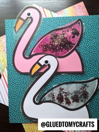 Two paper flamingo crafts with large googly eyes and sparkly glitter in their wings, placed on colorful patterned backgrounds—perfect inspiration if you love making a swan suncatcher. A watermark reads @GLUEDTOMYCRAFTS in the bottom right corner.