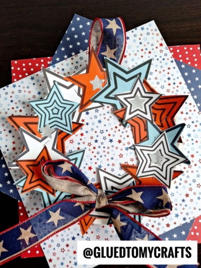 A star wreath made of layered star cutouts in red, blue, and black sits on star-patterned paper. It’s decorated with a blue ribbon with white stars and finished with a bow. The text @GLUEDTOMYCRAFTS appears at the bottom.