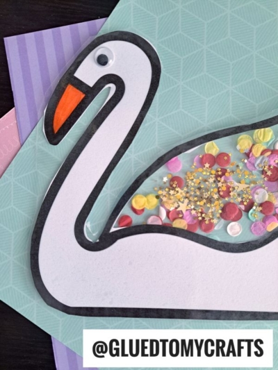 A swan suncatcher craft with a googly eye, orange beak, and a clear wing filled with colorful sequins and confetti, resting on pastel-colored paper layers. Text reads @GLUEDTOMYCRAFTS.