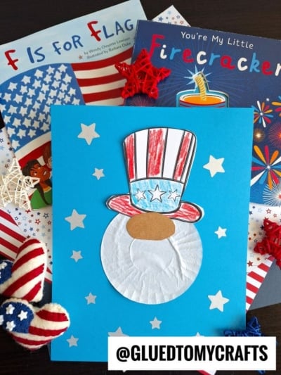 A children’s craft features a paper Uncle Sam face, resembling a cupcake liner gnome, made with a colored hat, white beard, and stars on blue paper. Patriotic-themed books and red, white, and blue decorations surround the craft. @GLUEDTOMYCRAFTS is at the bottom.