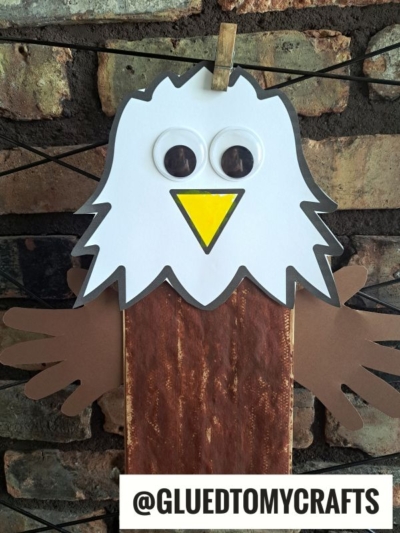 A handmade paper bag bald eagle craft with a white paper head, large googly eyes, a yellow beak, brown paper wings, and a brown painted body, clipped to a string in front of a brick wall. Text reads @GLUEDTOMYCRAFTS.