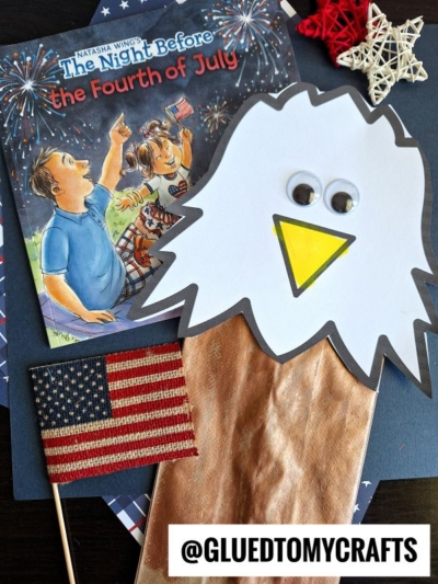 A children’s book titled “The Night Before the Fourth of July,” a paper bag bald eagle puppet, a small American flag, and star decorations are arranged on a table. The handle @GLUEDTOMYCRAFTS is at the bottom of the image.