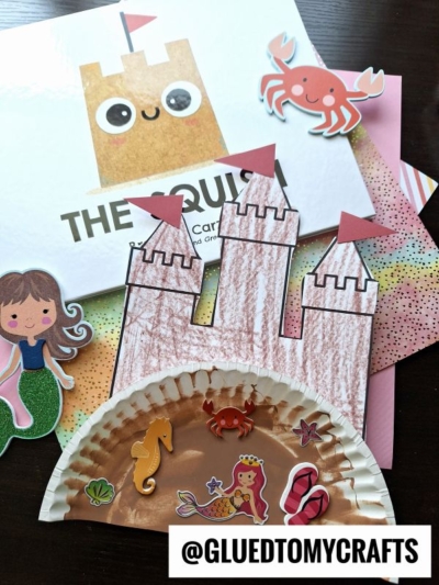 A paper plate sand castle is decorated and surrounded by cut-out stickers of a mermaid, crab, seahorse, and other sea-themed shapes. A book with a sandcastle illustration sits in the background. Text: @GLUEDTOMYCRAFTS.