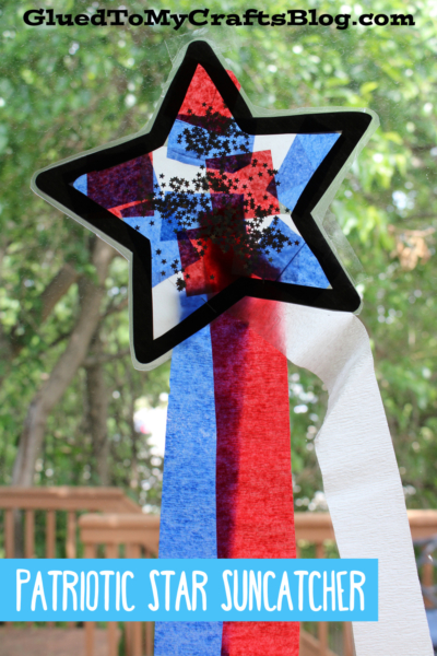 A patriotic star suncatcher with red, white, and blue tissue paper and black glitter hangs on a window. Red, white, and blue crepe paper streamers dangle below. Trees and a wooden deck can be seen outside.