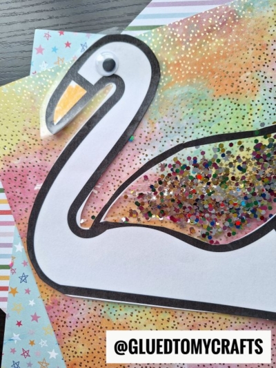 A swan suncatcher craft made on colorful star-patterned paper, decorated with a googly eye, outlined in black, and filled with multicolored glitter. The handle @GLUEDTOMYCRAFTS appears at the bottom.