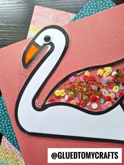 A handmade swan suncatcher craft featuring a white paper outline, a googly eye, and colorful sequins inside the body, placed on layered polka dot papers. The Instagram handle @GLUEDTOMYCRAFTS is visible.
