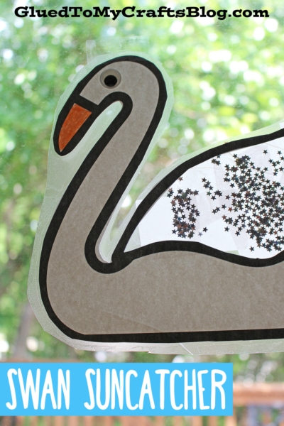 A handmade swan suncatcher craft with a gray outline and orange beak, decorated with silver star glitter, shines on a window. The text reads Swan Suncatcher and GluedToMyCraftsBlog.com. Perfect for your next swan suncatcher project!.