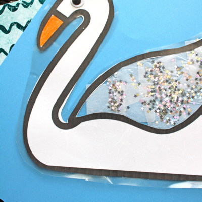 A swan suncatcher craft featuring a white paper body, orange beak, and a clear wing filled with shiny star-shaped glitter, set against a blue background. The text reads SWAN SUNCATCHER.
