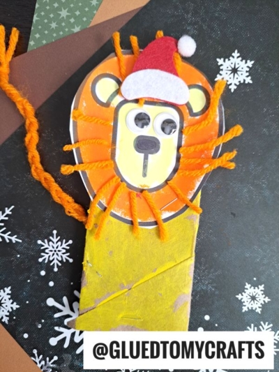 A handmade toilet paper roll lion with a yellow face, orange yarn mane, googly eyes, and a red Santa hat sits on a painted tube against a black background with white snowflakes. Text: @GLUEDTOMYCRAFTS.