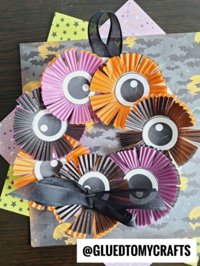 A festive cupcake liner Halloween wreath made of orange, black, and purple cupcake liners with paper eyes, arranged on Halloween-themed paper. A black ribbon is tied on the bottom. The handle @GLUEDTOMYCRAFTS appears on the bottom right.